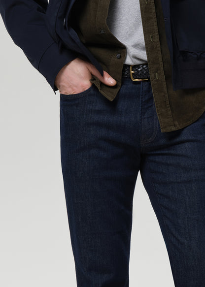 Close-up of a person wearing dark blue denim jeans, a gray t-shirt, an olive green overshirt, and a navy blazer, with one hand in the pocket of the jeans, against a light gray background.