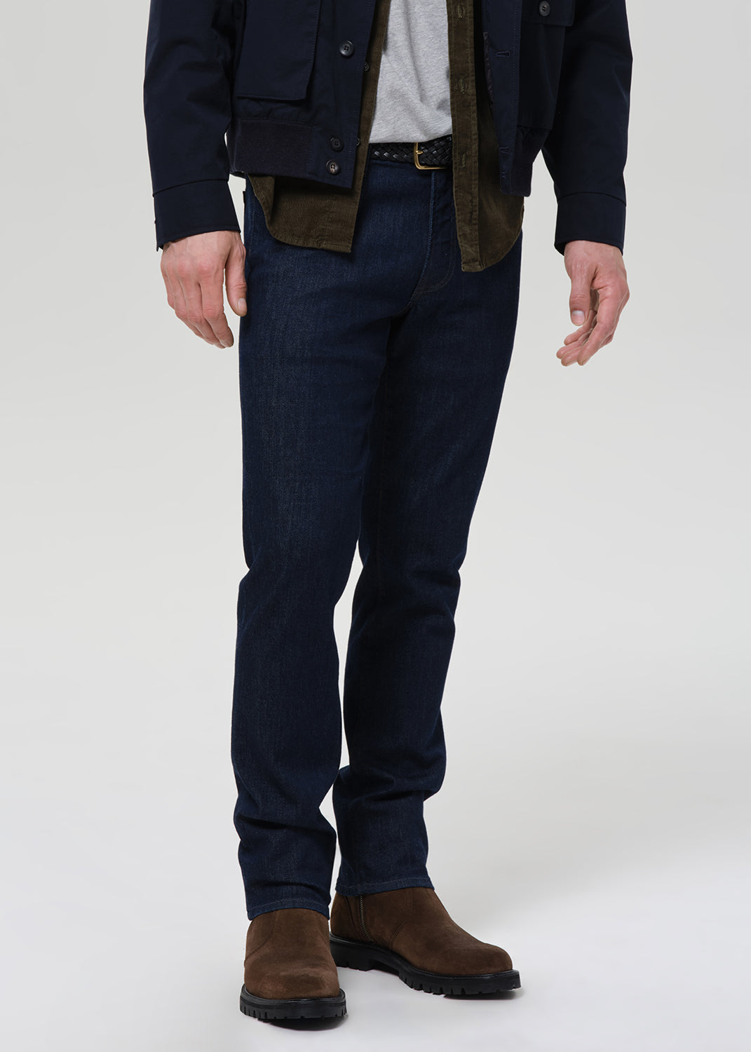 A person shown from the chest down wearing dark navy blue jeans, a dark jacket, a light gray shirt, and brown suede boots, photographed against a plain light gray studio background.