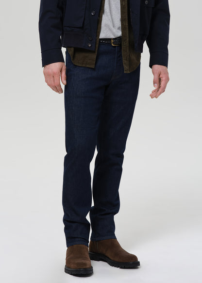 A person shown from the chest down wearing dark navy blue jeans, a dark jacket, a light gray shirt, and brown suede boots, photographed against a plain light gray studio background.