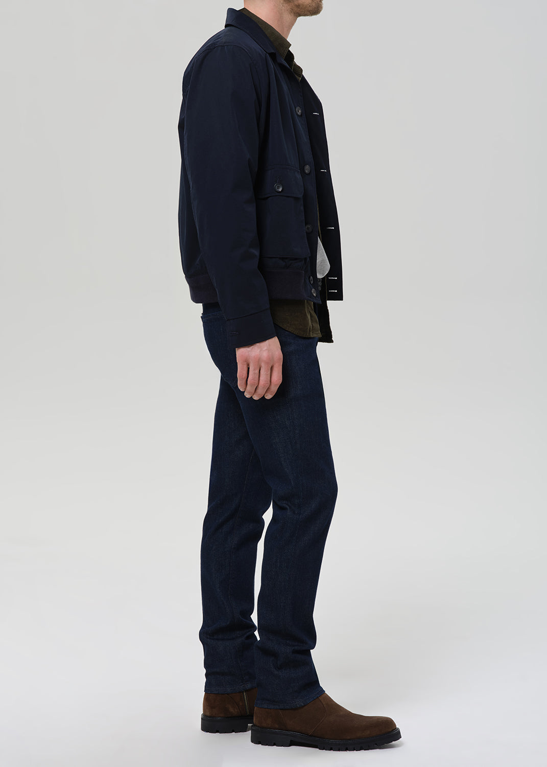 A man shown in profile wearing a dark navy jacket, matching dark navy trousers, and brown boots against a light gray studio background.
