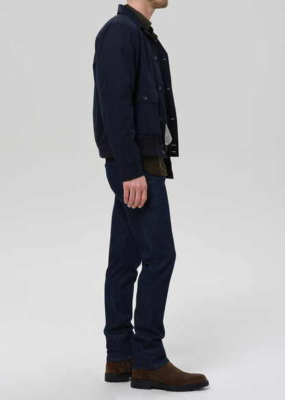 A man shown in profile wearing a dark navy jacket, matching dark navy trousers, and brown boots against a light gray studio background.