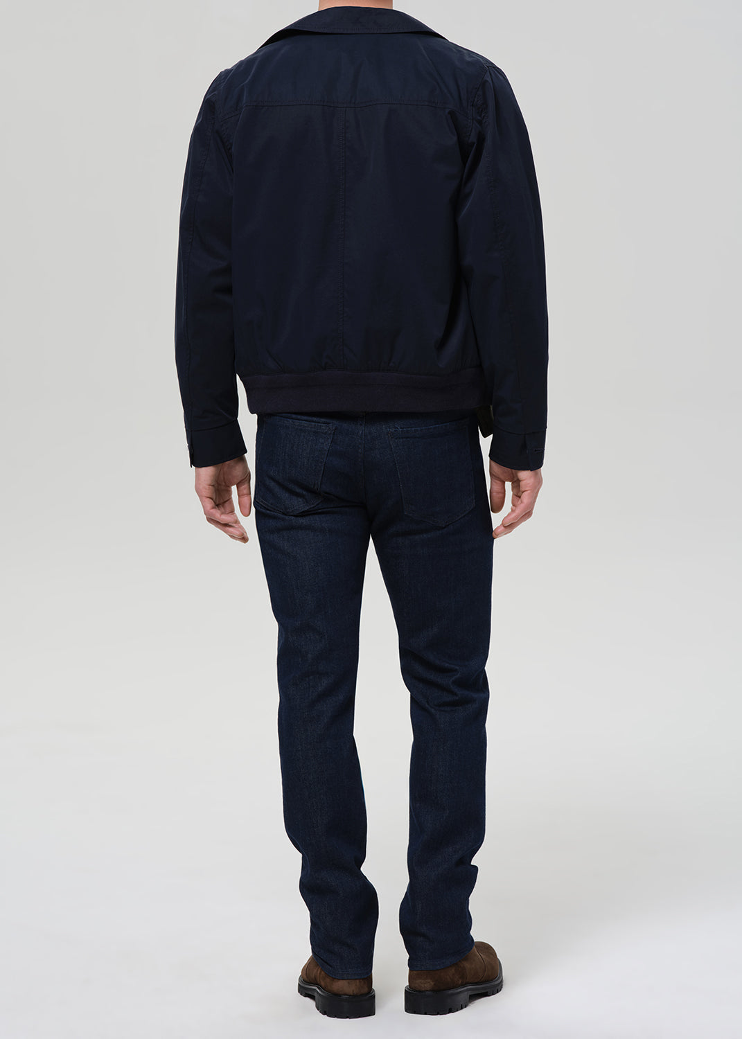 A man viewed from the back wearing a dark navy jacket and dark blue jeans, standing against a light gray studio background.
