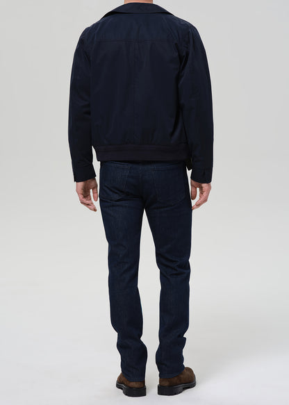 A man viewed from the back wearing a dark navy jacket and dark blue jeans, standing against a light gray studio background.