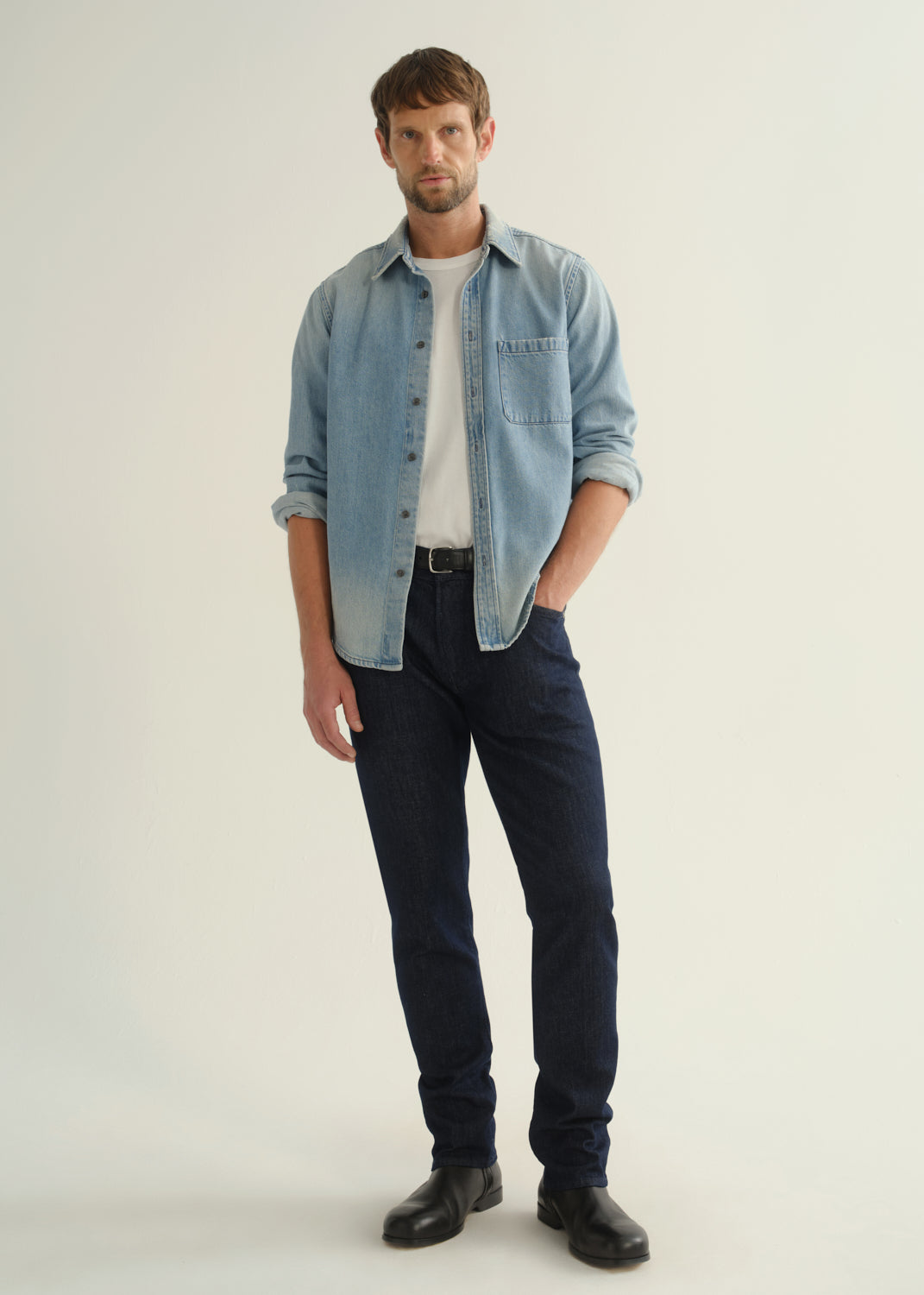 Person wearing a light blue denim shirt, dark indigo jeans, and black boots, front full body view on a light grey background.