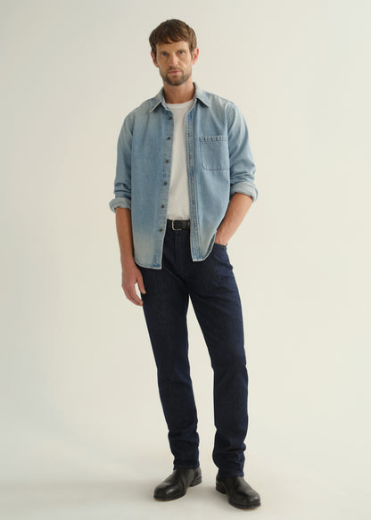 Person wearing a light blue denim shirt, dark indigo jeans, and black boots, front full body view on a light grey background.