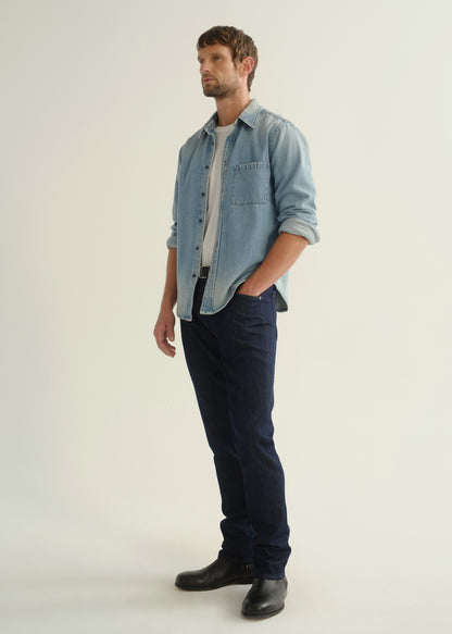 Person wearing a light blue denim shirt, dark indigo jeans, and black boots, angled front full body view on a light grey background.