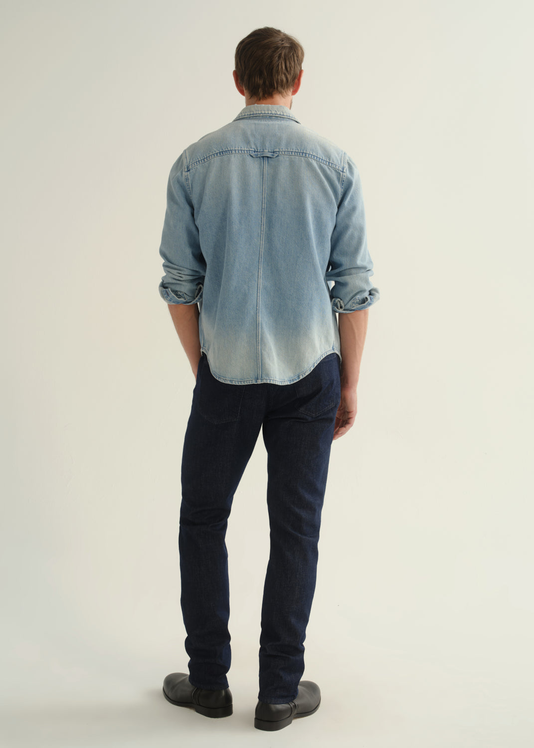 Person wearing a light blue denim shirt, dark indigo jeans, and black boots, back full body view on a light grey background.