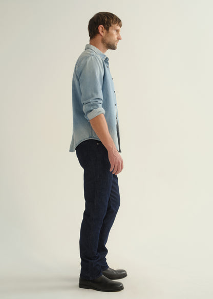 Person wearing a light blue denim shirt, dark indigo jeans, and black boots, side full body view on a light grey background.