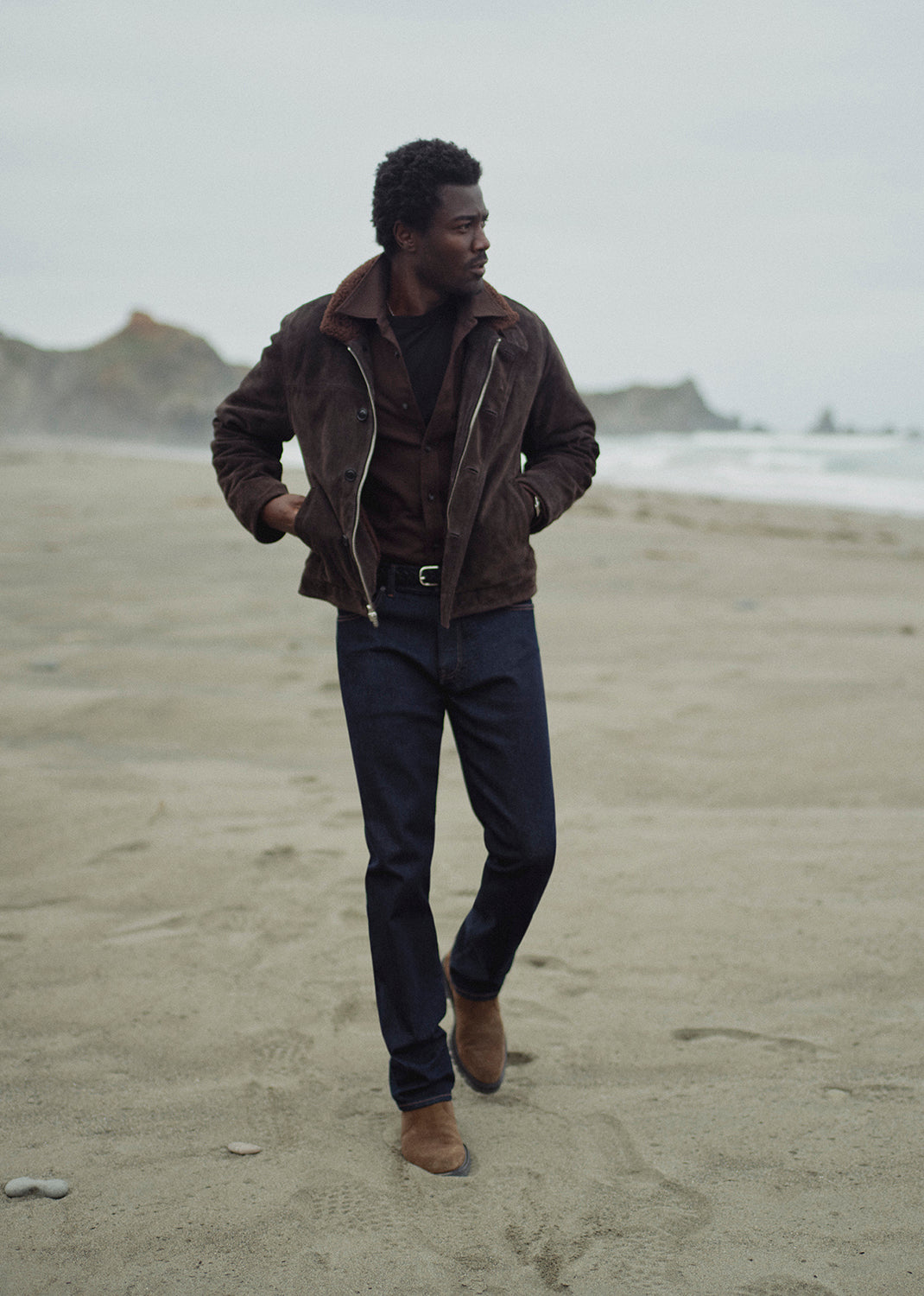 A man walking on a sandy beach wearing a brown suede jacket, dark blue jeans, and brown boots under an overcast sky.