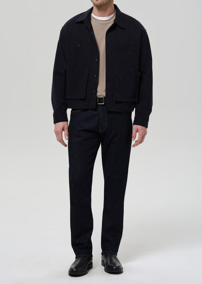 Person wearing a black jacket, beige shirt, and black pants on a grey background