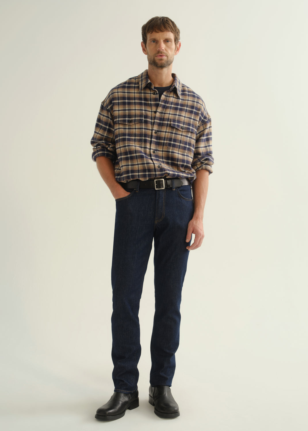 Person wearing a brown and navy plaid shirt, dark indigo jeans, and black boots, front full body view on a light grey background.