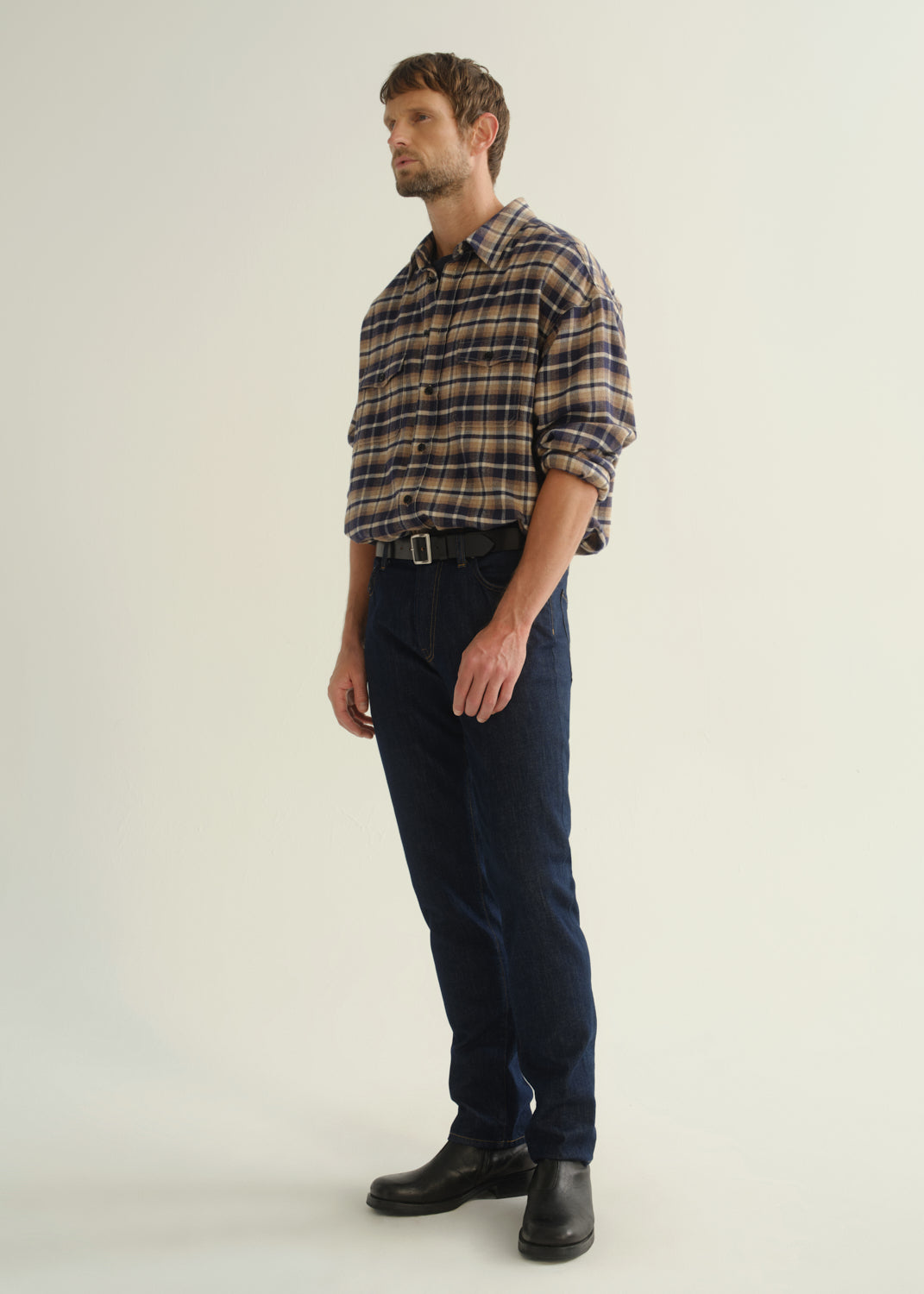 Person wearing a brown and navy plaid shirt, dark indigo jeans, and black boots, angled front full body view on a light grey background.