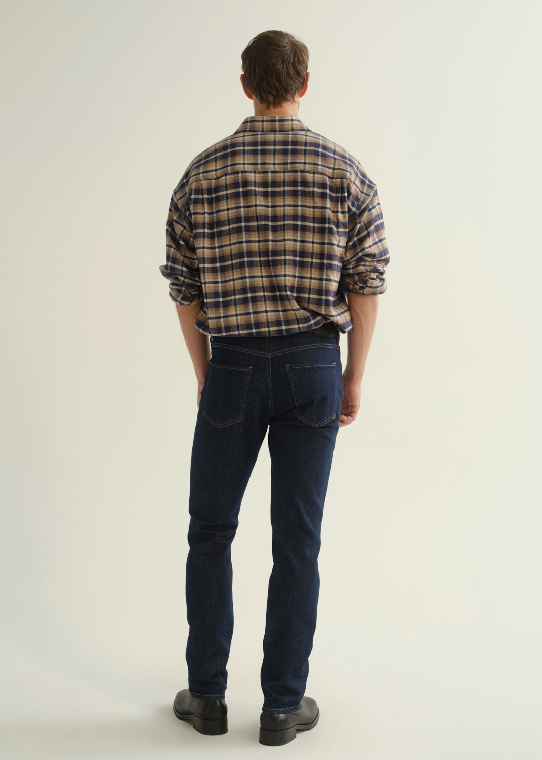 Person wearing a brown and navy plaid shirt, dark indigo jeans, and black boots, back full body view on a light grey background.