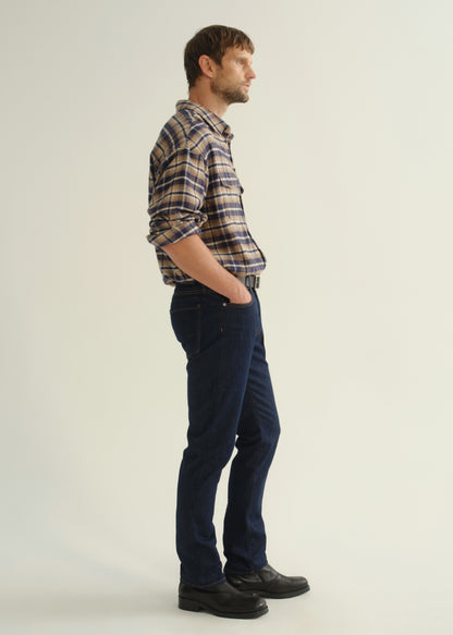 Person wearing a brown and navy plaid shirt, dark indigo jeans, and black boots, side full body view on a light grey background.
