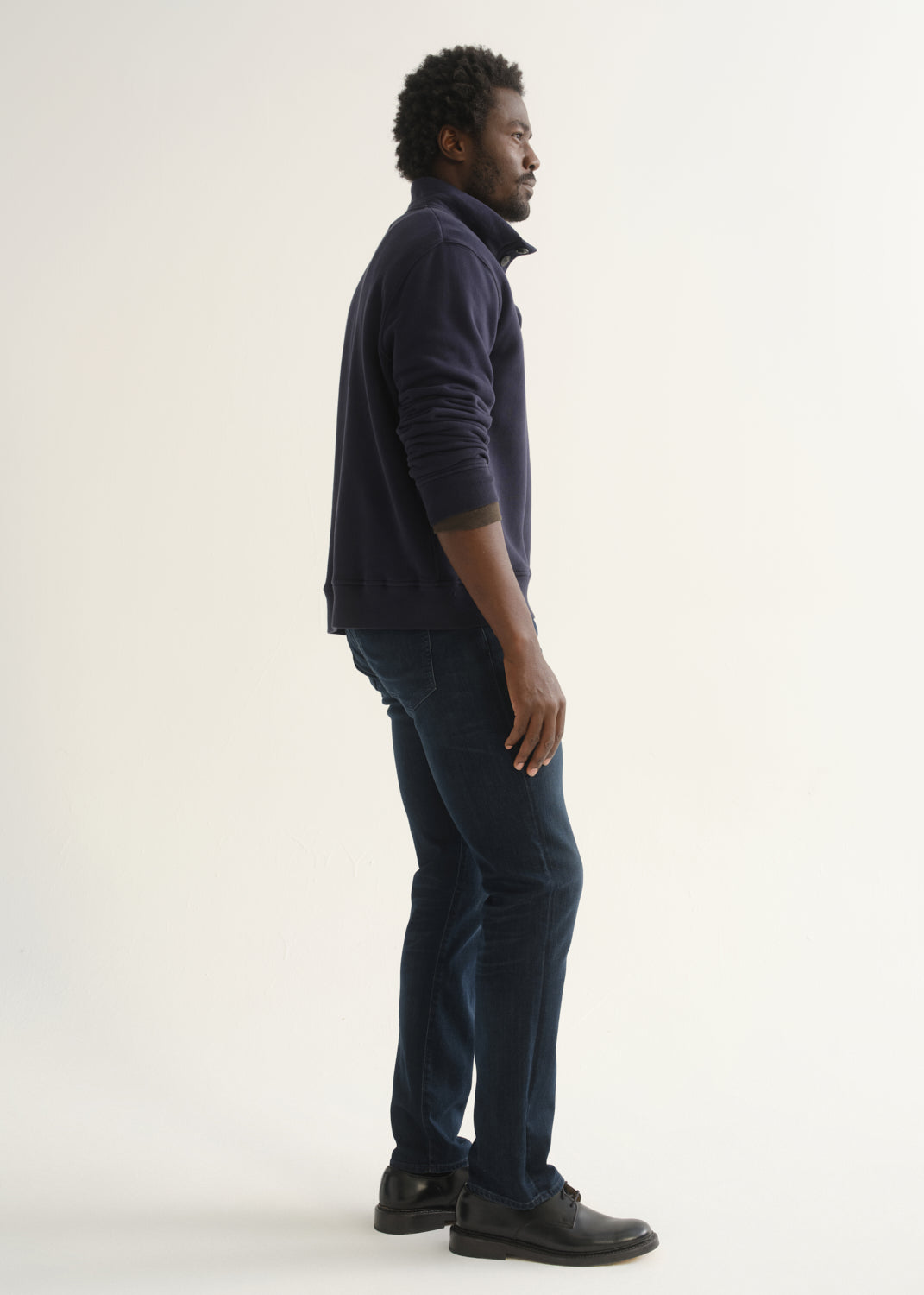 Person wearing a dark navy long sleeve shirt, dark indigo jeans, and black boots, side full body view on a light grey background.