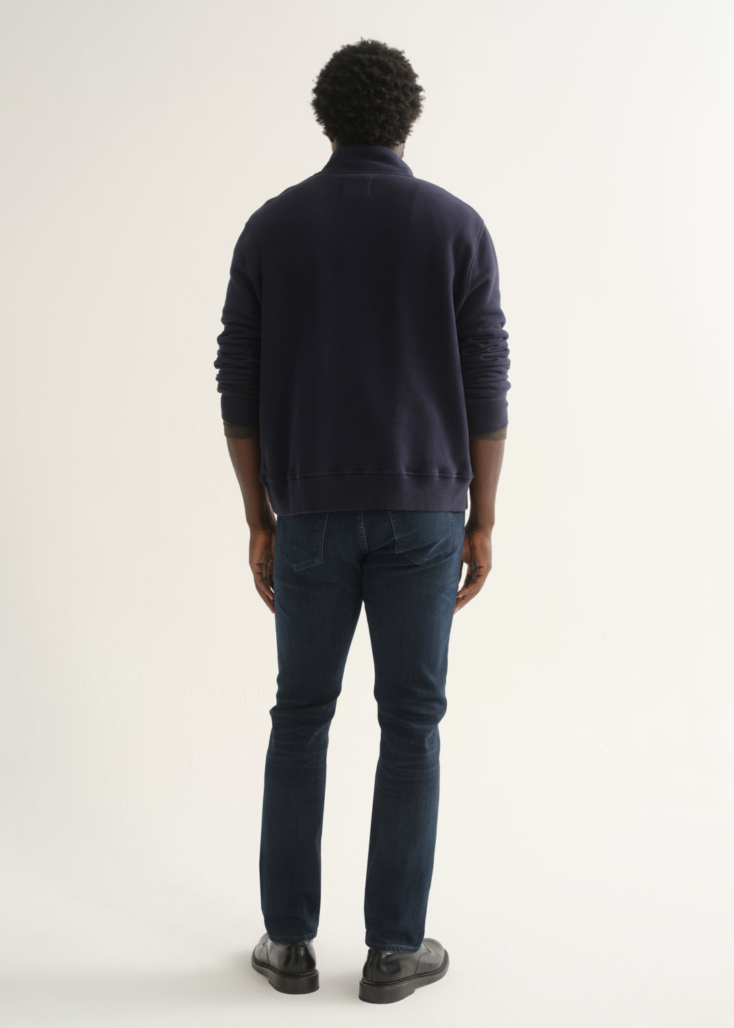 Person wearing a dark navy long sleeve shirt, dark indigo jeans, and black boots, back full body view on a light grey background.