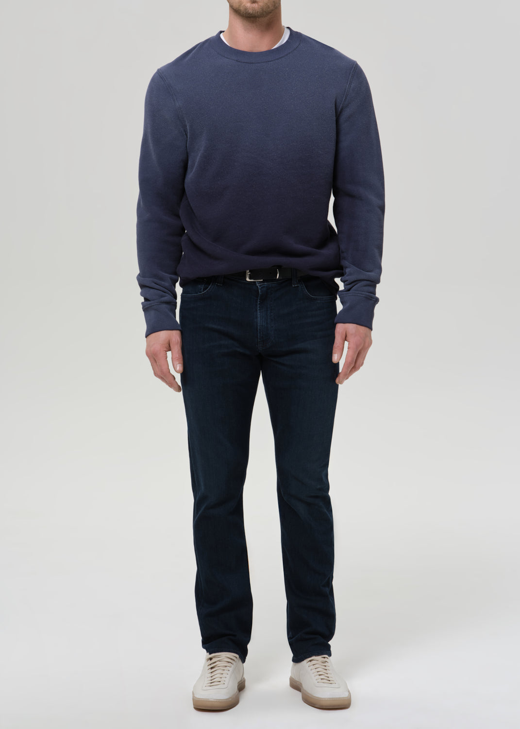 A man wearing a navy blue crewneck sweatshirt, dark blue jeans, and beige sneakers, standing against a grey background.