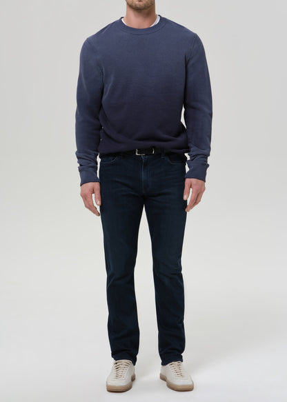 A man wearing a navy blue crewneck sweatshirt, dark blue jeans, and beige sneakers, standing against a grey background.