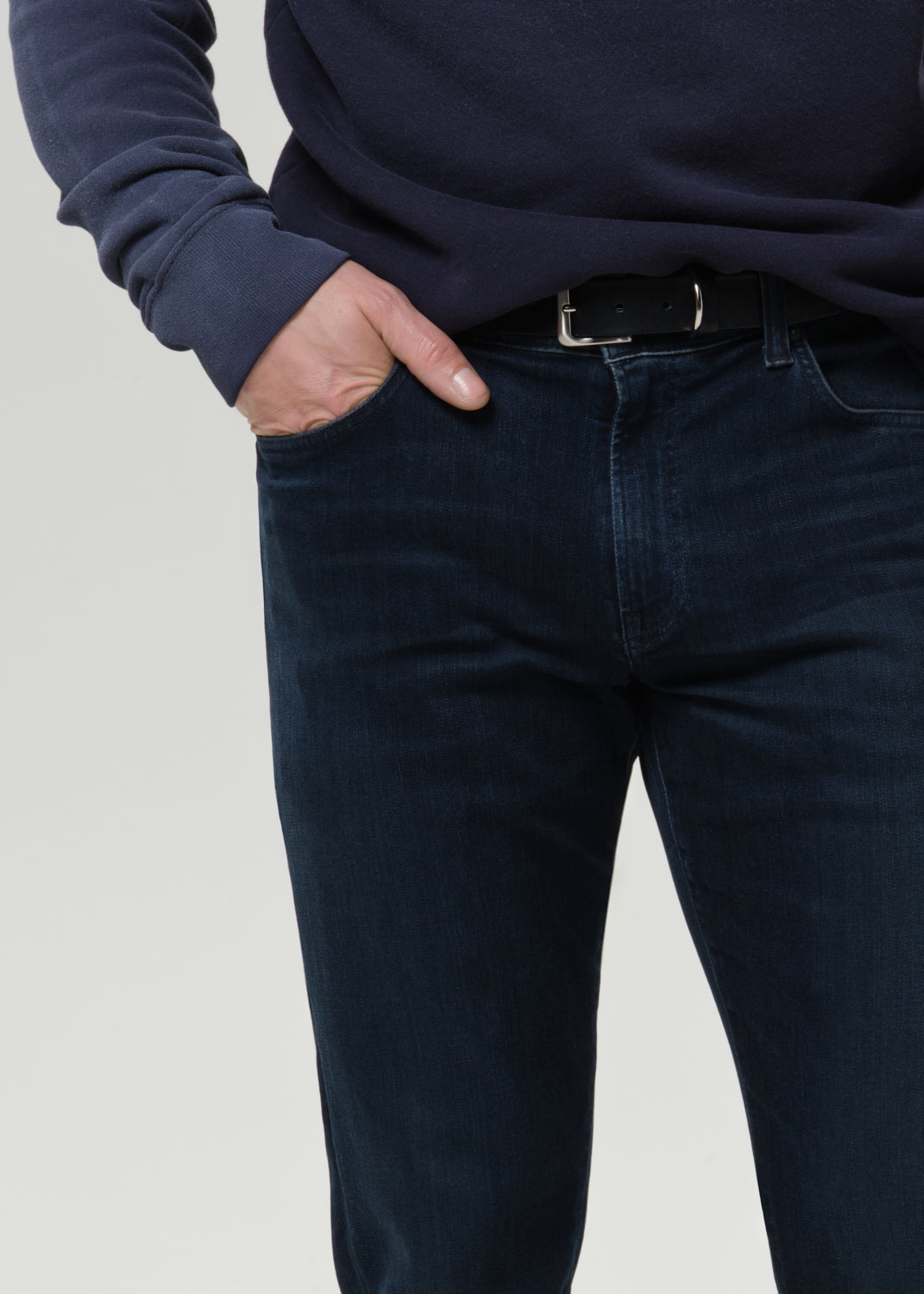 A close-up of a person wearing dark navy blue jeans with a black belt and a matching dark navy long-sleeve top, standing against a grey background.