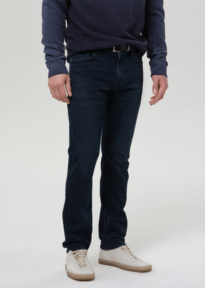 A person wearing dark navy slim-fit jeans, a navy long-sleeve top, and off-white sneakers, shown from the chest down against a grey background.