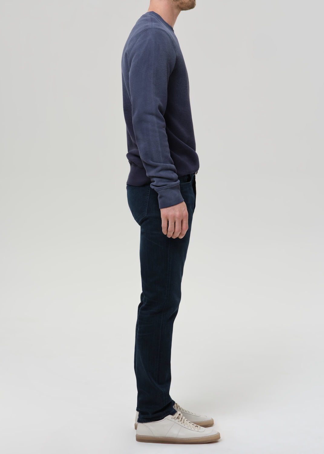 A man shown in profile wearing a dark navy long-sleeve knit top, dark blue jeans, and off-white sneakers against a grey background.