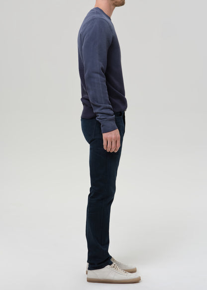 A man shown in profile wearing a dark navy long-sleeve knit top, dark blue jeans, and off-white sneakers against a grey background.