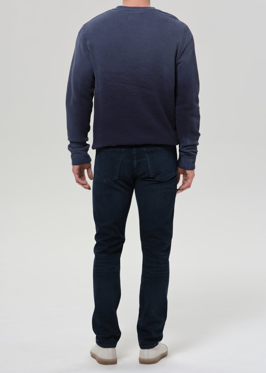 A man viewed from the back wearing a dark navy blue long-sleeve crewneck sweatshirt and dark blue jeans, standing against a grey background.