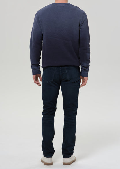 A man viewed from the back wearing a dark navy blue long-sleeve crewneck sweatshirt and dark blue jeans, standing against a grey background.