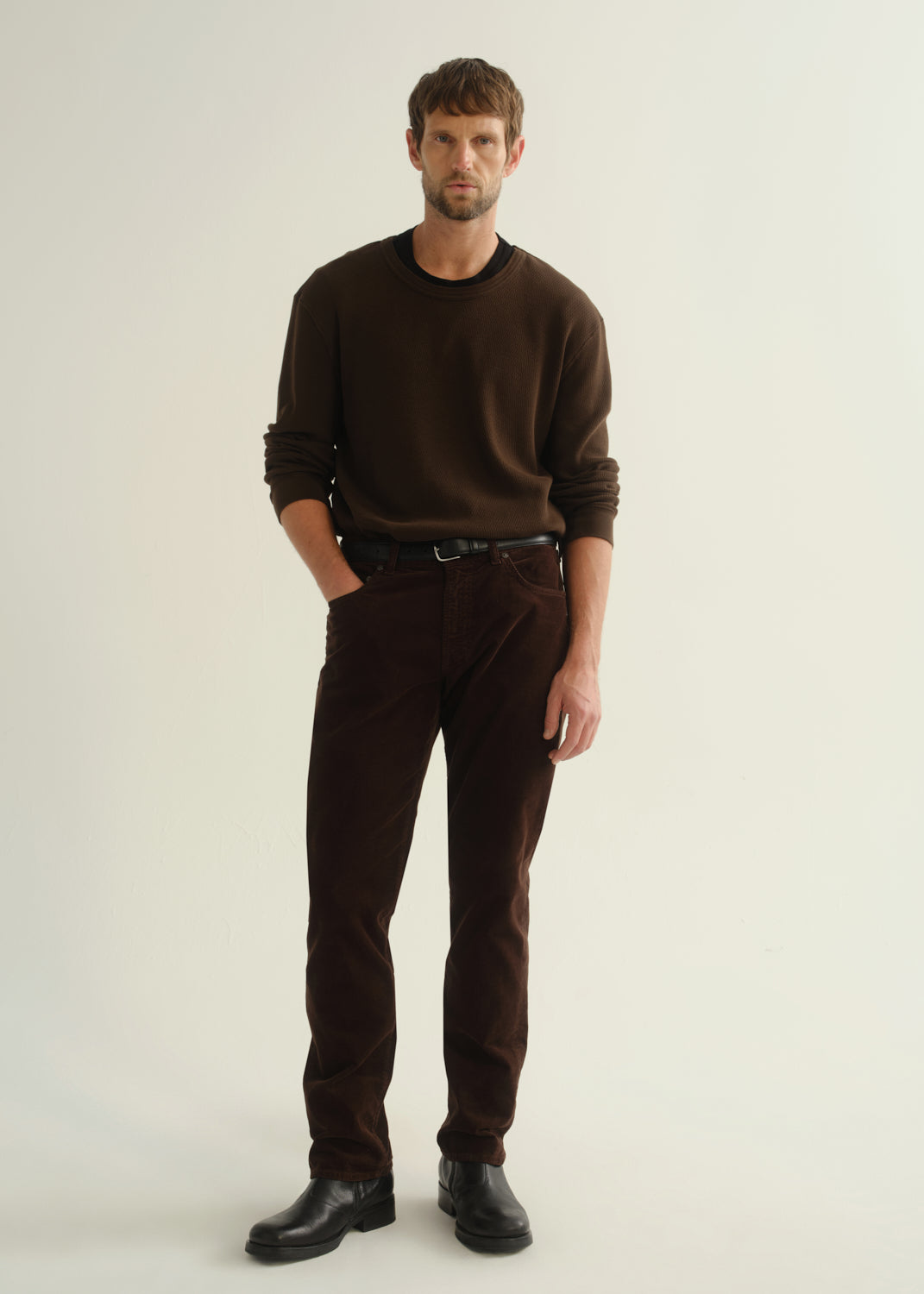 Person wearing a dark brown crewneck sweater, dark brown pants, and black boots, front full body view on a light grey background.