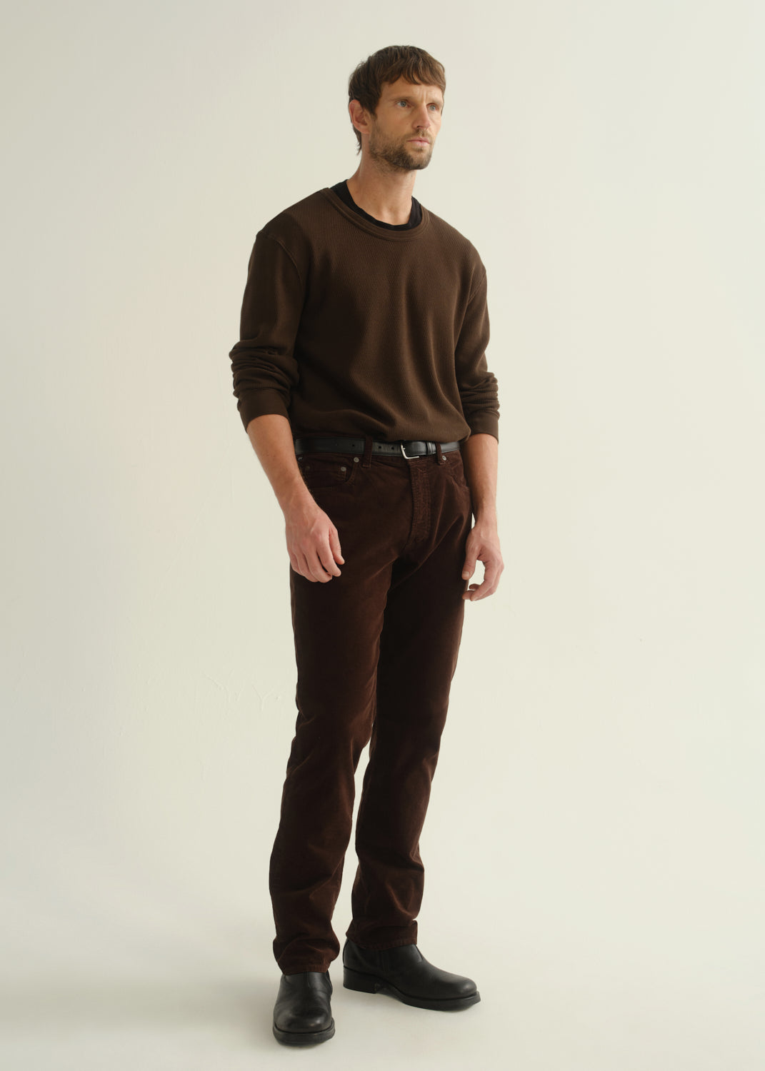 Person wearing a dark brown crewneck sweater, dark brown pants, and black boots, angled front full body view on a light grey background.