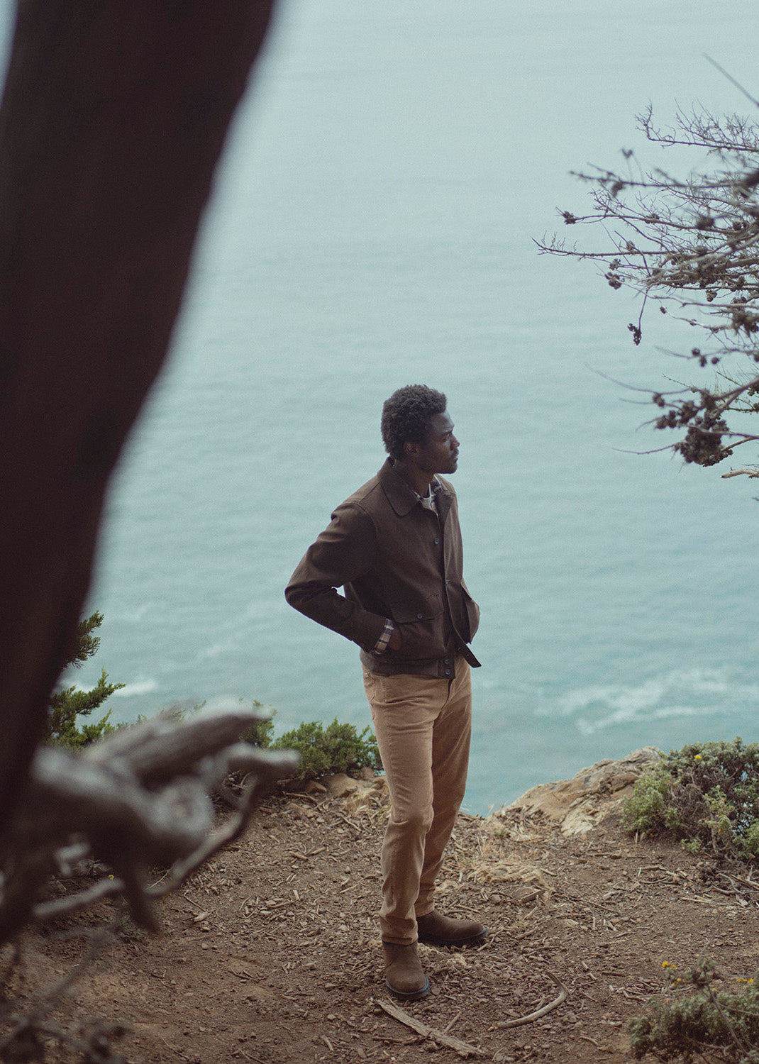 A man stands on a rocky cliffside overlooking the ocean, wearing a brown jacket, tan pants, and brown boots. The scene is framed by blurred tree branches in the foreground, with the sea and sky in the background.