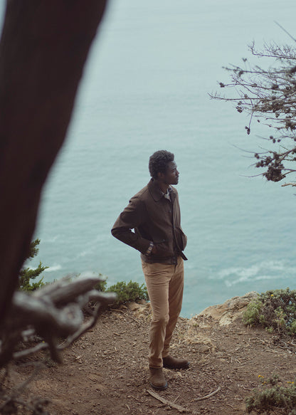 A man stands on a rocky cliffside overlooking the ocean, wearing a brown jacket, tan pants, and brown boots. The scene is framed by blurred tree branches in the foreground, with the sea and sky in the background.