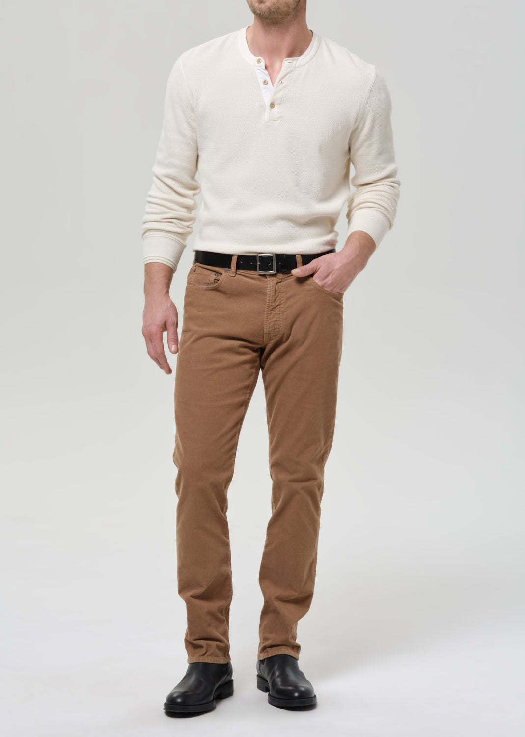 Man wearing a cream sweater, brown pants, and black shoes on a grey background