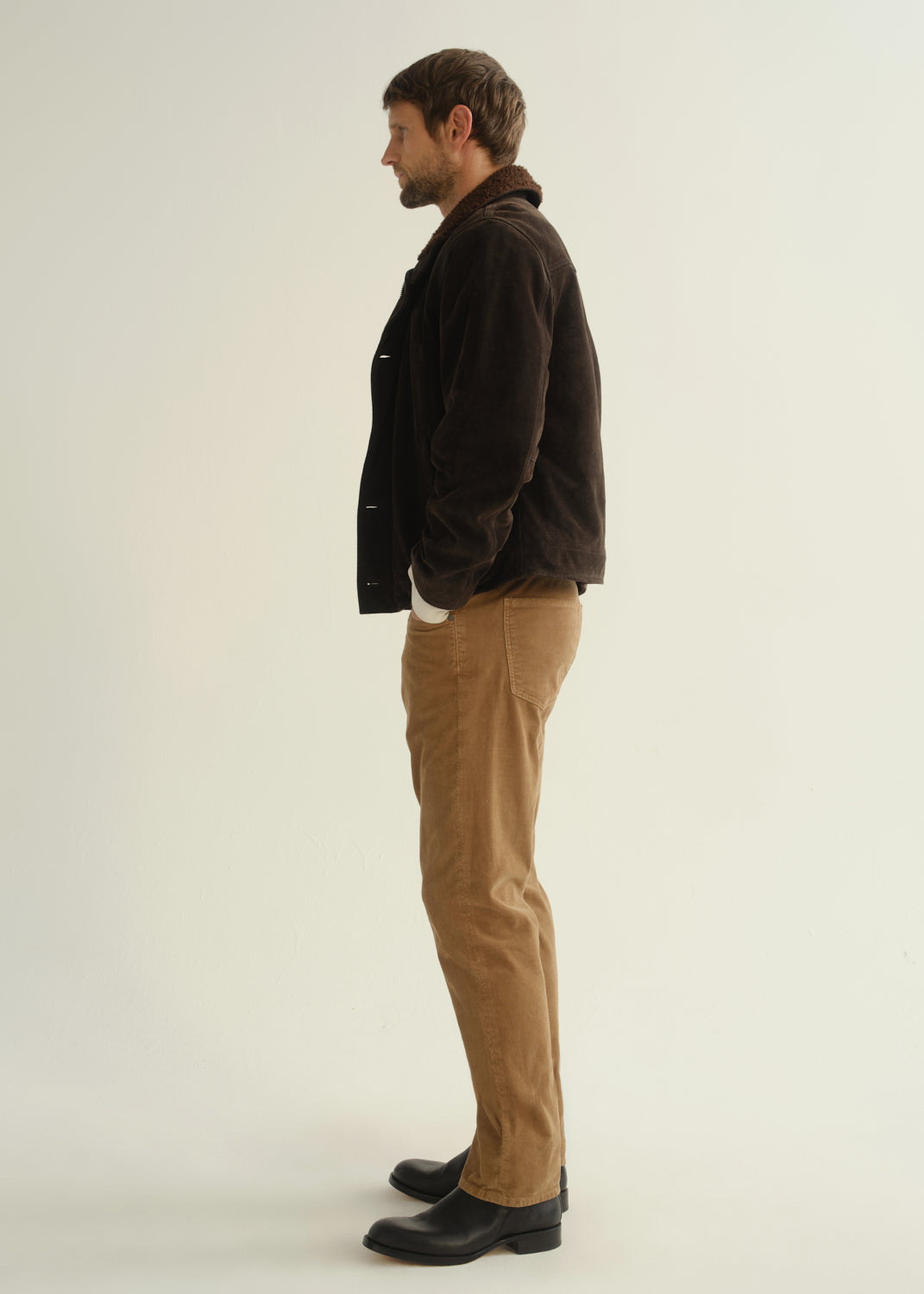 Person wearing a dark brown jacket, black shirt, tan corduroy pants, and black boots, side full body view on a light grey background.