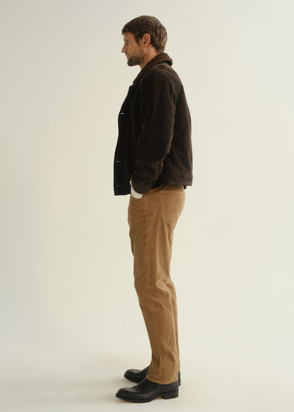 Person wearing a dark brown jacket, black shirt, tan corduroy pants, and black boots, side full body view on a light grey background.