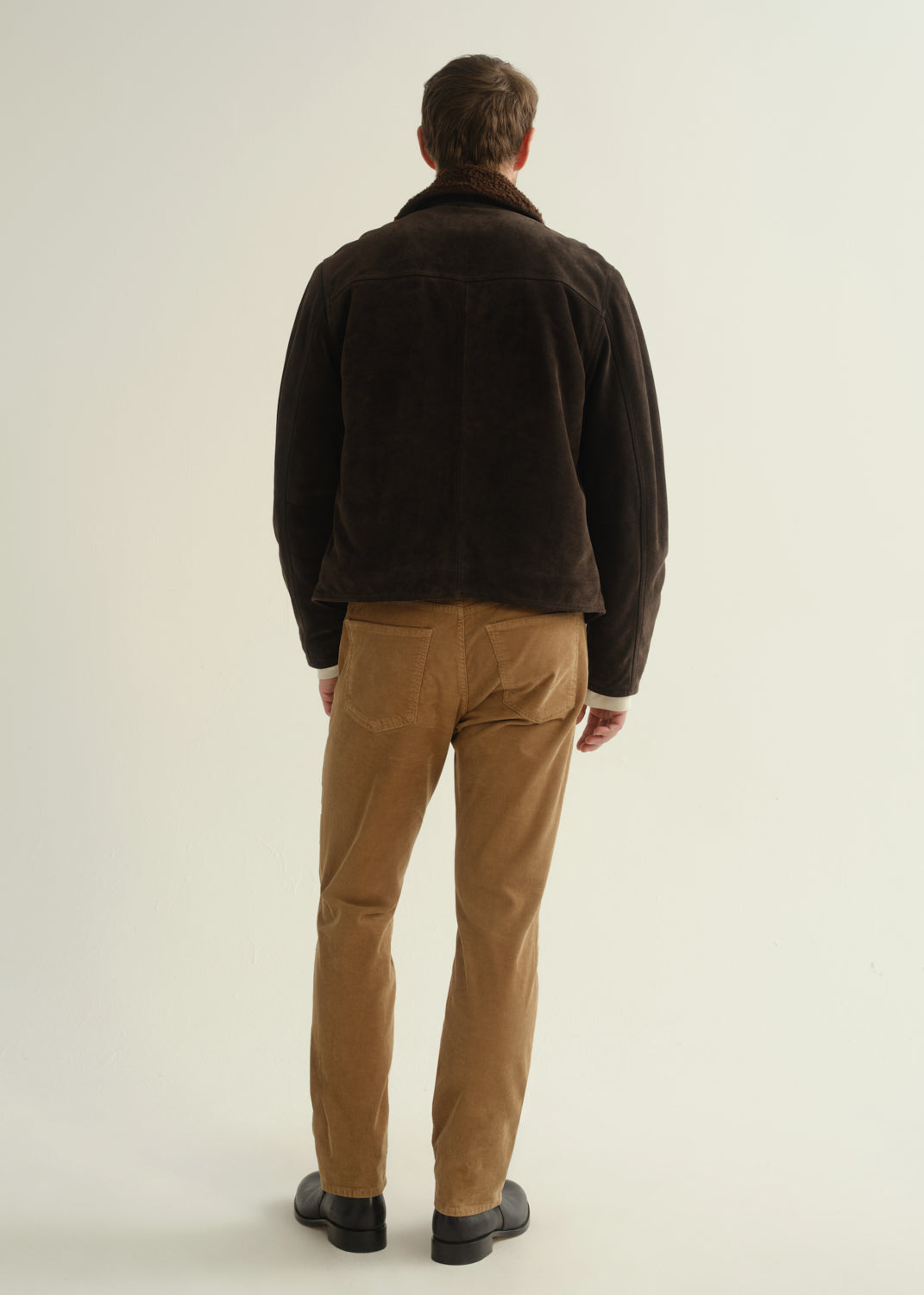 Person wearing a dark brown jacket, black shirt, tan corduroy pants, and black boots, back full body view on a light grey background.