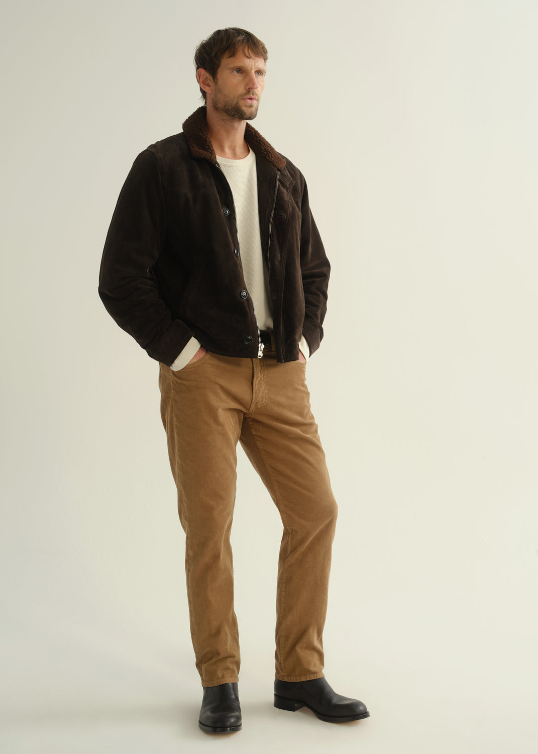 Person wearing a dark brown jacket, black shirt, tan corduroy pants, and black boots, angled front full body view on a light grey background.