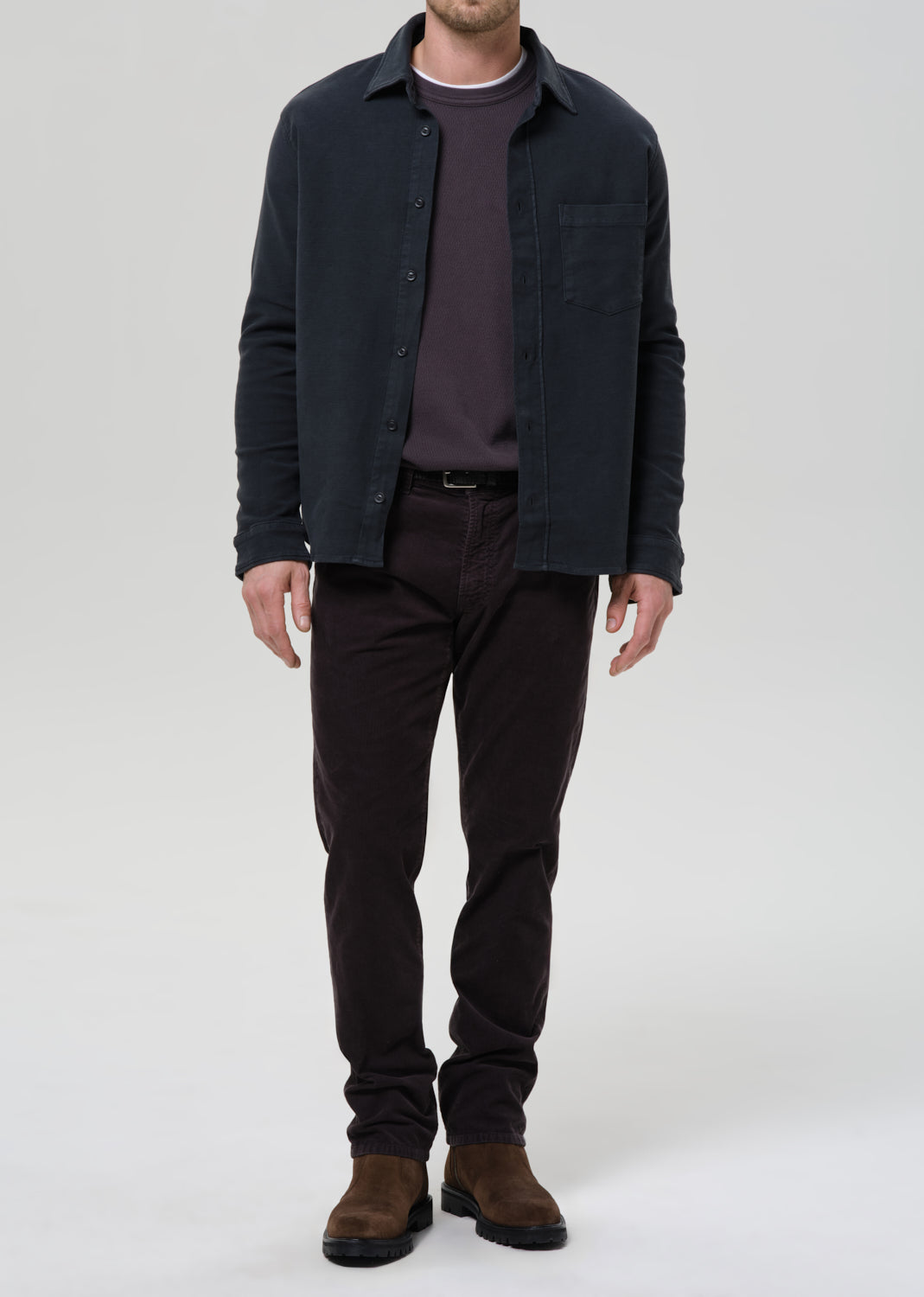 Man wearing a dark jacket, shirt, pants, and shoes on a grey background
