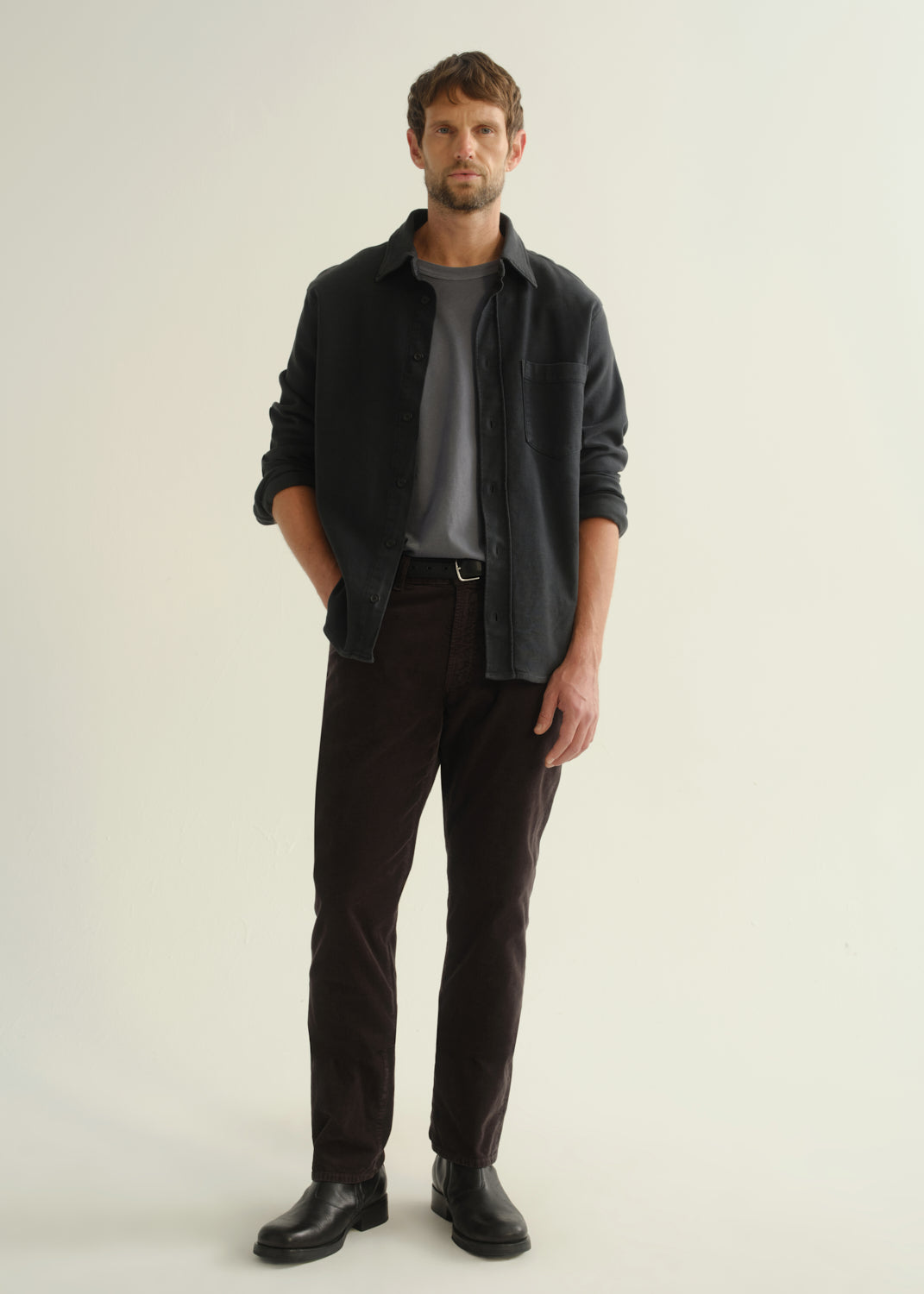Person wearing a dark navy button-front shirt over a grey top, washed black jeans, and black boots, front full body view on a light grey background.