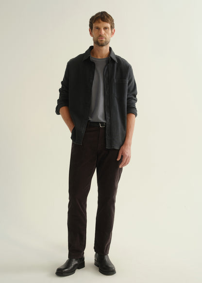 Person wearing a dark navy button-front shirt over a grey top, washed black jeans, and black boots, front full body view on a light grey background.