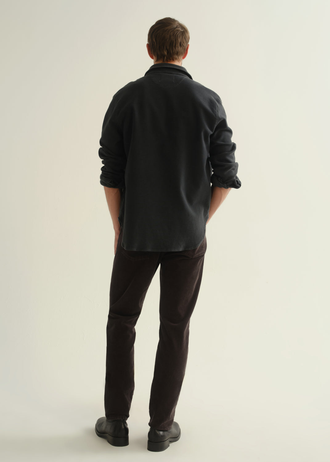Person wearing a dark navy button-front shirt, washed black jeans, and black boots, back full body view on a light grey background.