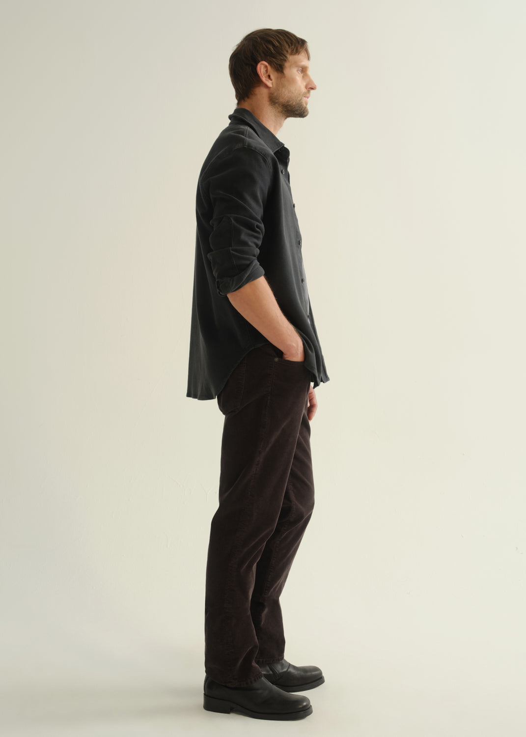 Person wearing a dark navy button-front shirt, washed black jeans, and black boots, side full body view on a light grey background.