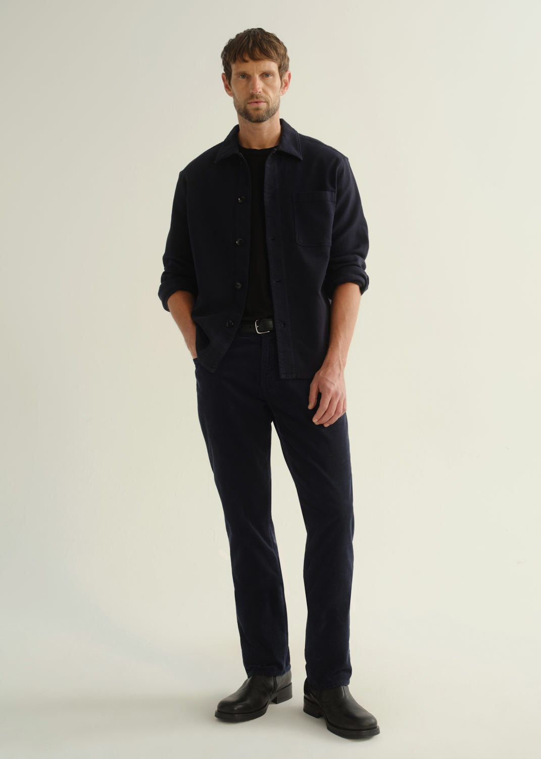 Person wearing a dark navy button-front shirt, dark indigo jeans, and black boots, front full body view on a light grey background.