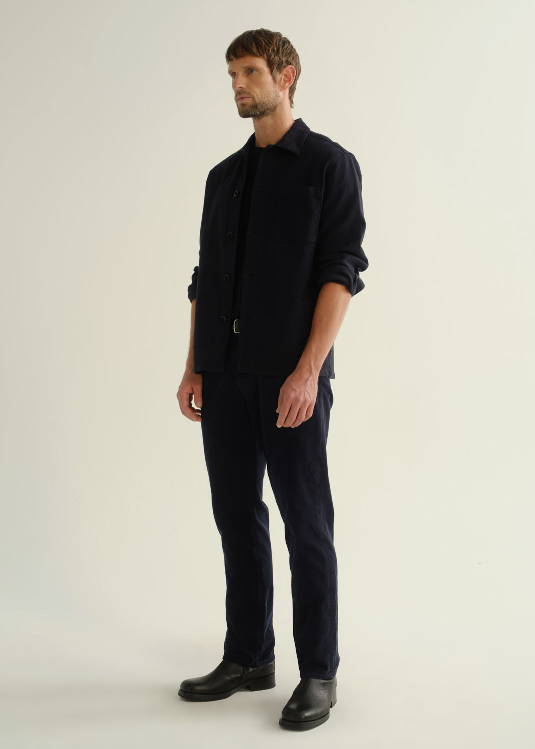 Person wearing a dark navy button-front shirt, dark indigo jeans, and black boots, angled front full body view on a light grey background.