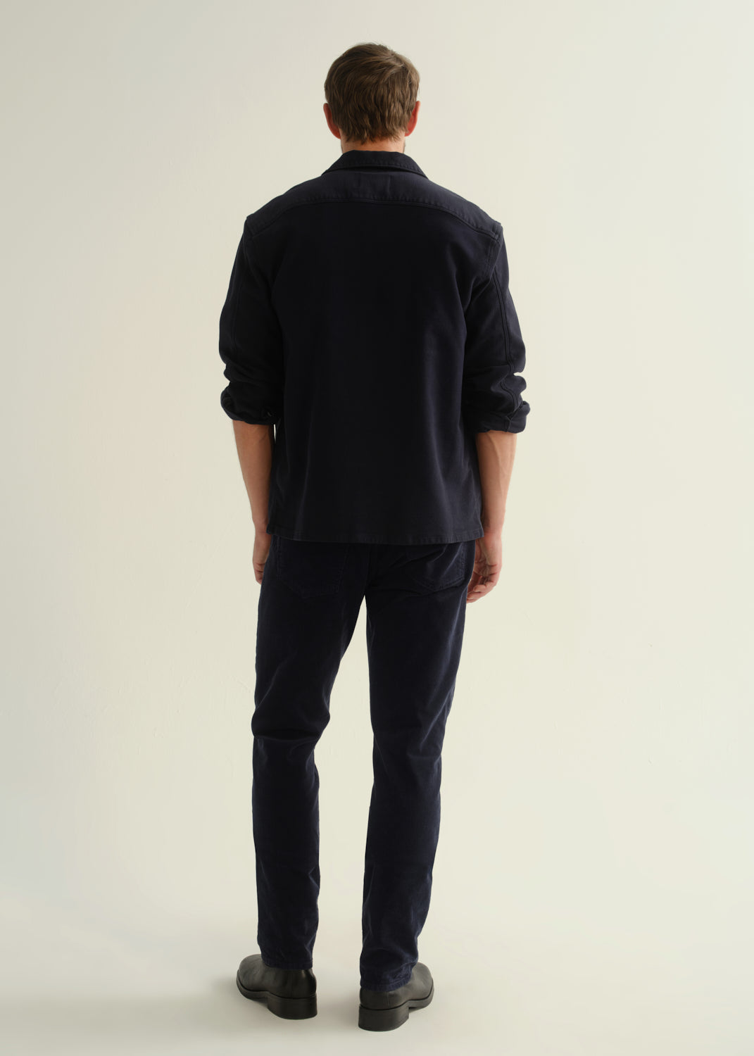 Person wearing a dark navy button-front shirt, dark indigo jeans, and black boots, back full body view on a light grey background.