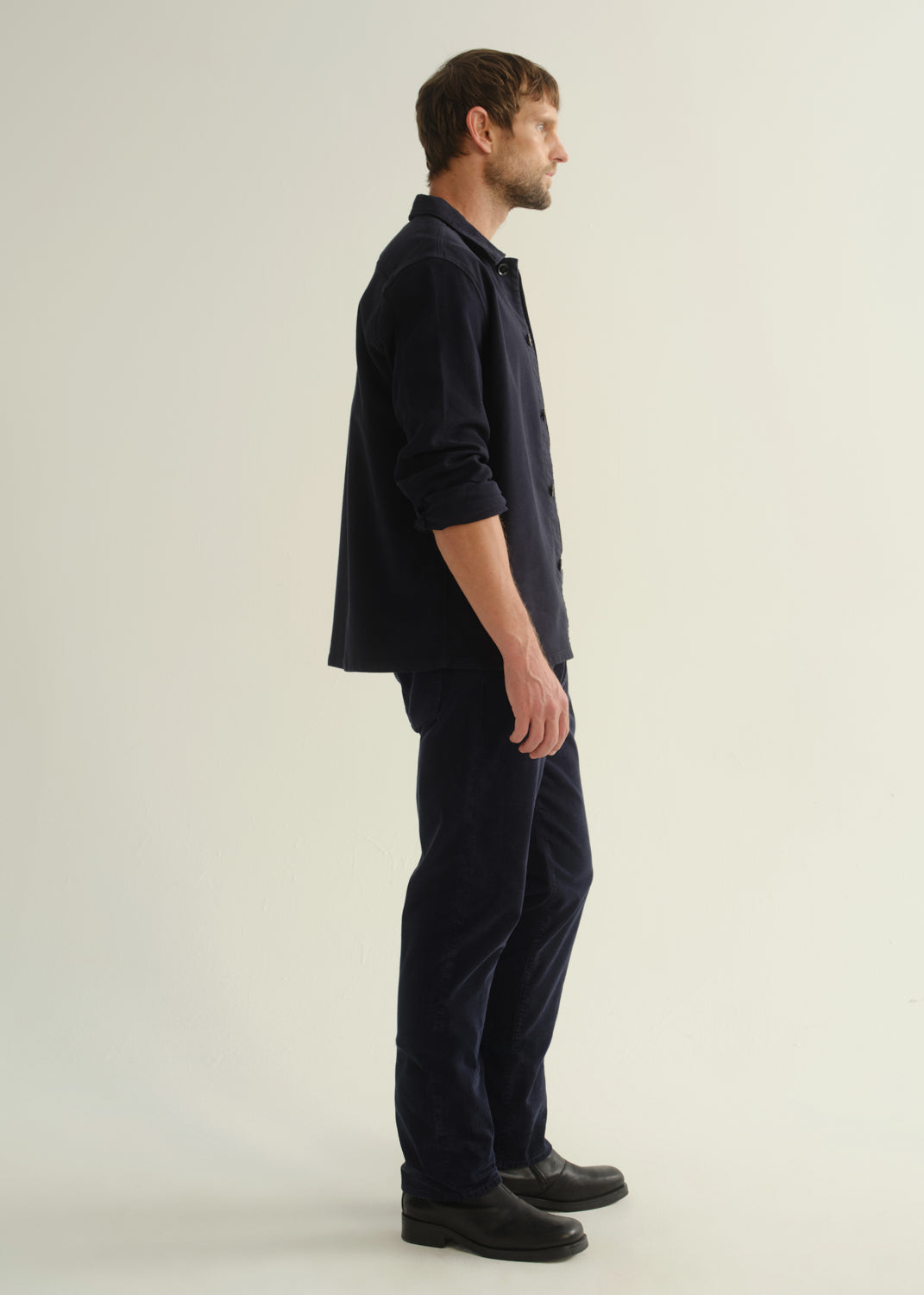 Person wearing a dark navy button-front shirt, dark indigo jeans, and black boots, side full body view on a light grey background.