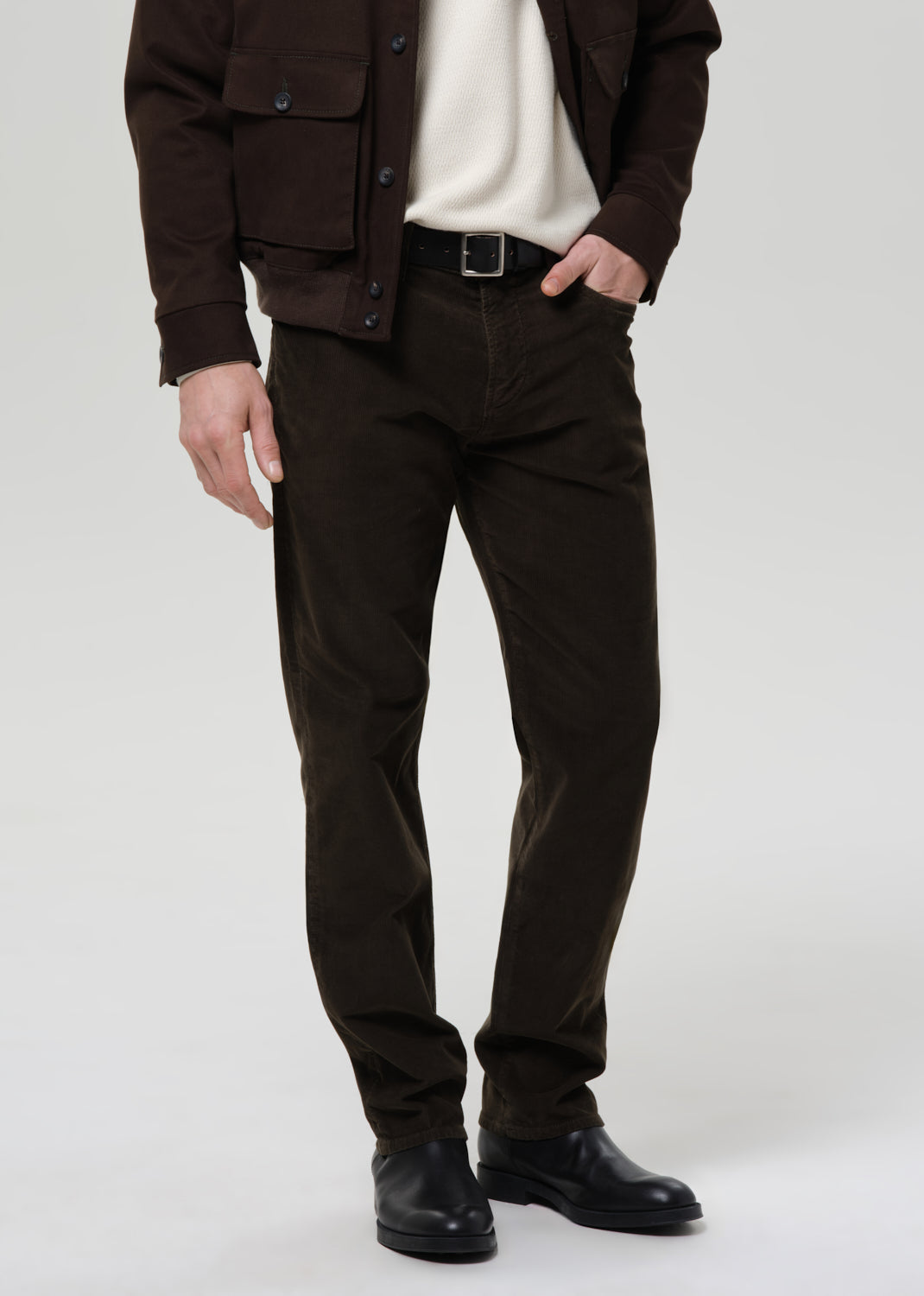 Person wearing a brown jacket, white shirt, dark green pants, and black shoes on a grey background