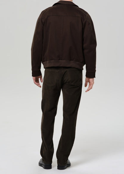 Person wearing a brown jacket and dark green pants on a grey background