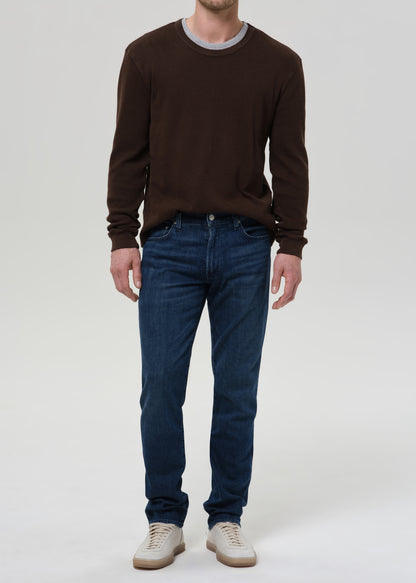 A man viewed from the front wearing a dark brown long-sleeve henley shirt and dark blue jeans, standing against a grey background.