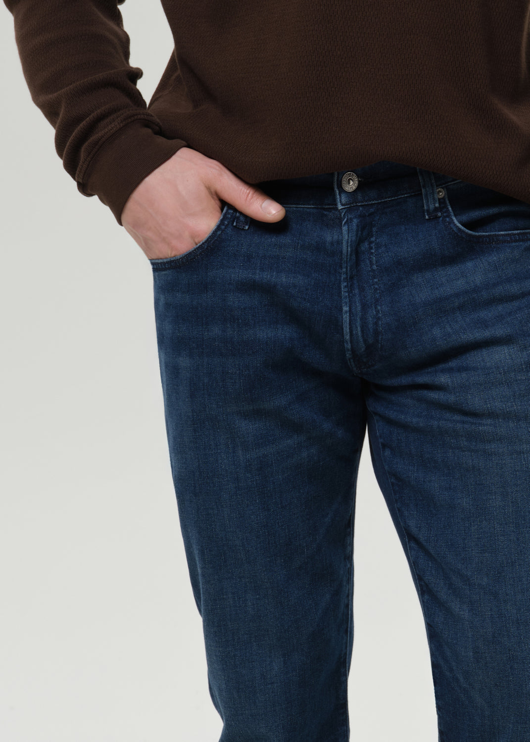 A close-up of a person wearing dark blue denim jeans and a brown ribbed sweater, with one hand in the pocket, against a grey background.